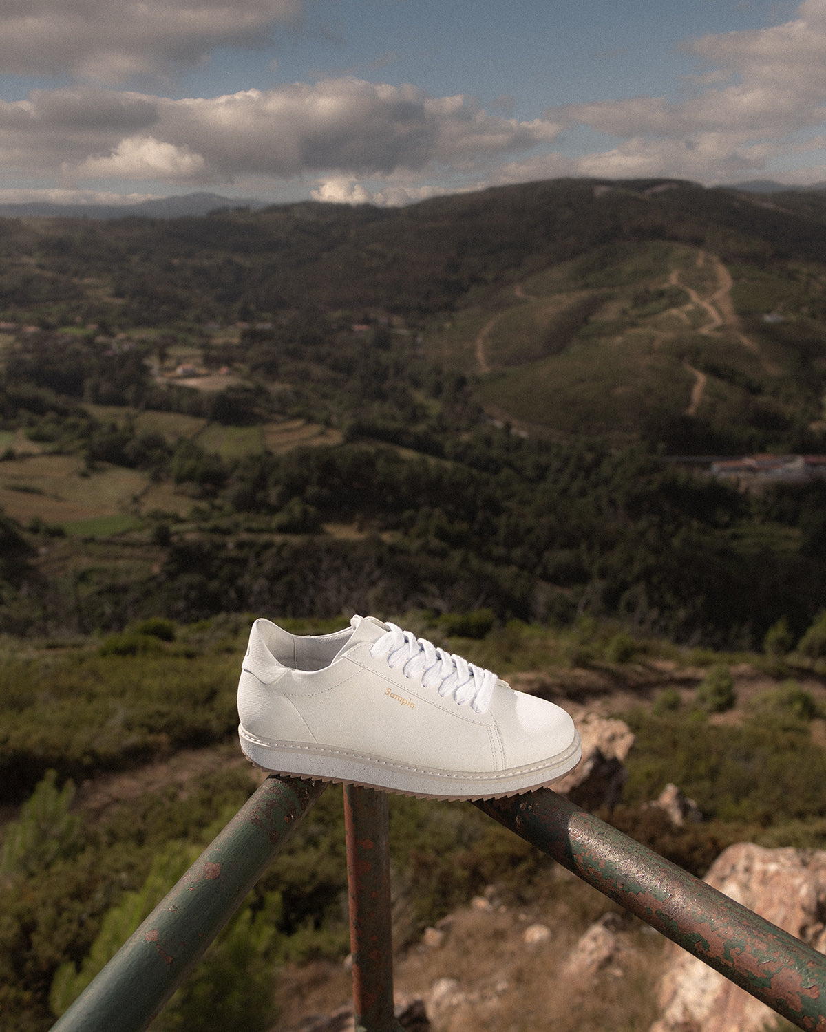 White sneaker on a metal railing with a scenic view of hills and clouds in the background